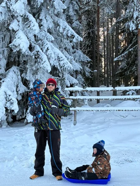 Isä kantaa pientä lasta sylissään lumisessa metsässä, toinen poika istuu pulkassa vieressä